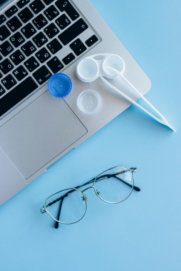 Flat lay of eyeglasses, contact lens case, and laptop on a blue surface.
